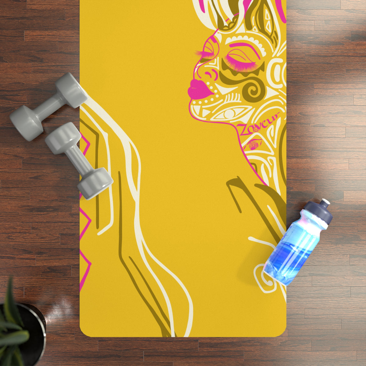 Top-down view of yellow Queen of Phoenix yoga mat with pink line-art face profile on a wood floor beside dumbbells