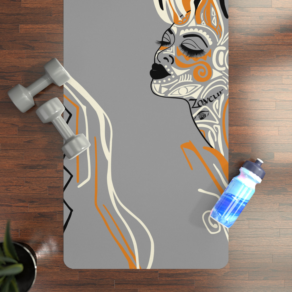 Top-down view of grey Queen of Phoenix yoga mat with illustrated face profile on a wood floor beside dumbbells