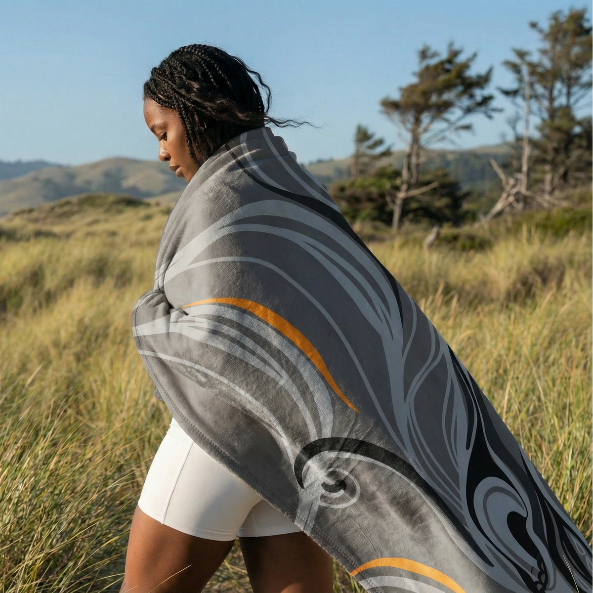 Woman walking through tall grass wrapped in the grey Queen of Phoenix blanket with flowing black and cream lines