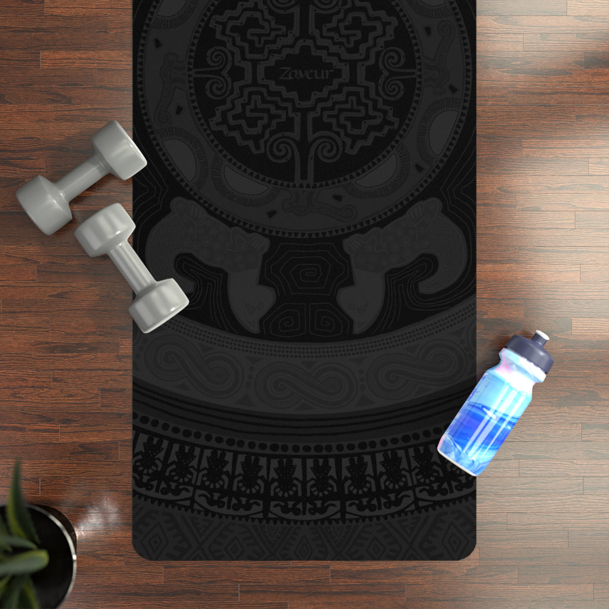 Top-down view of the Colombian Dreams black mandala yoga mat on a wood floor with dumbbells and a water bottle