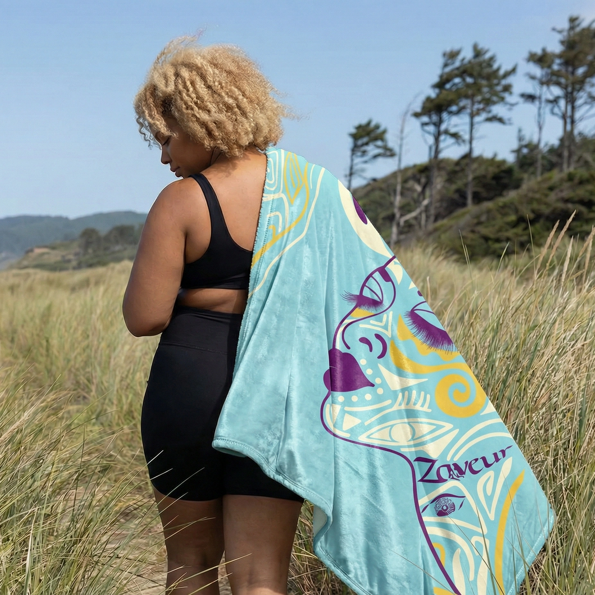 Woman on a coastal path with the aqua Queen of Phoenix blanket over one shoulder, the line-art face visible