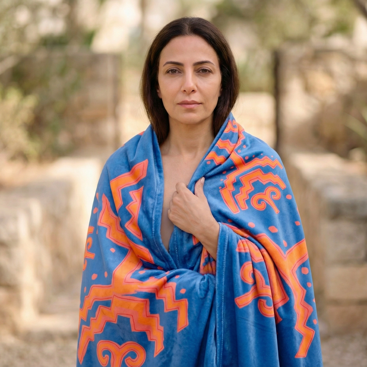 Woman wrapped in a blue Colombian Dreams blanket with bold orange geometric shapes, stone walls behind her