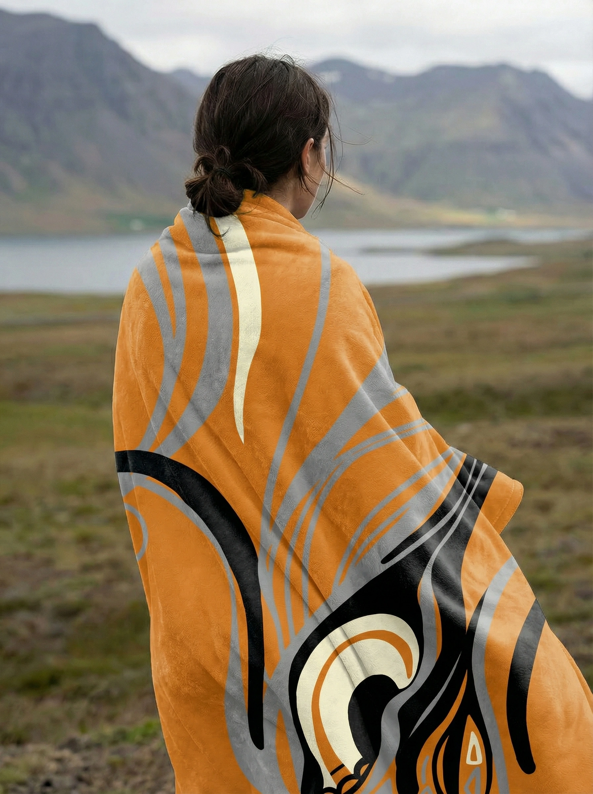 Woman seen from behind wrapped in the orange Queen of Phoenix blanket, flowing grey and black lines, lake and hills