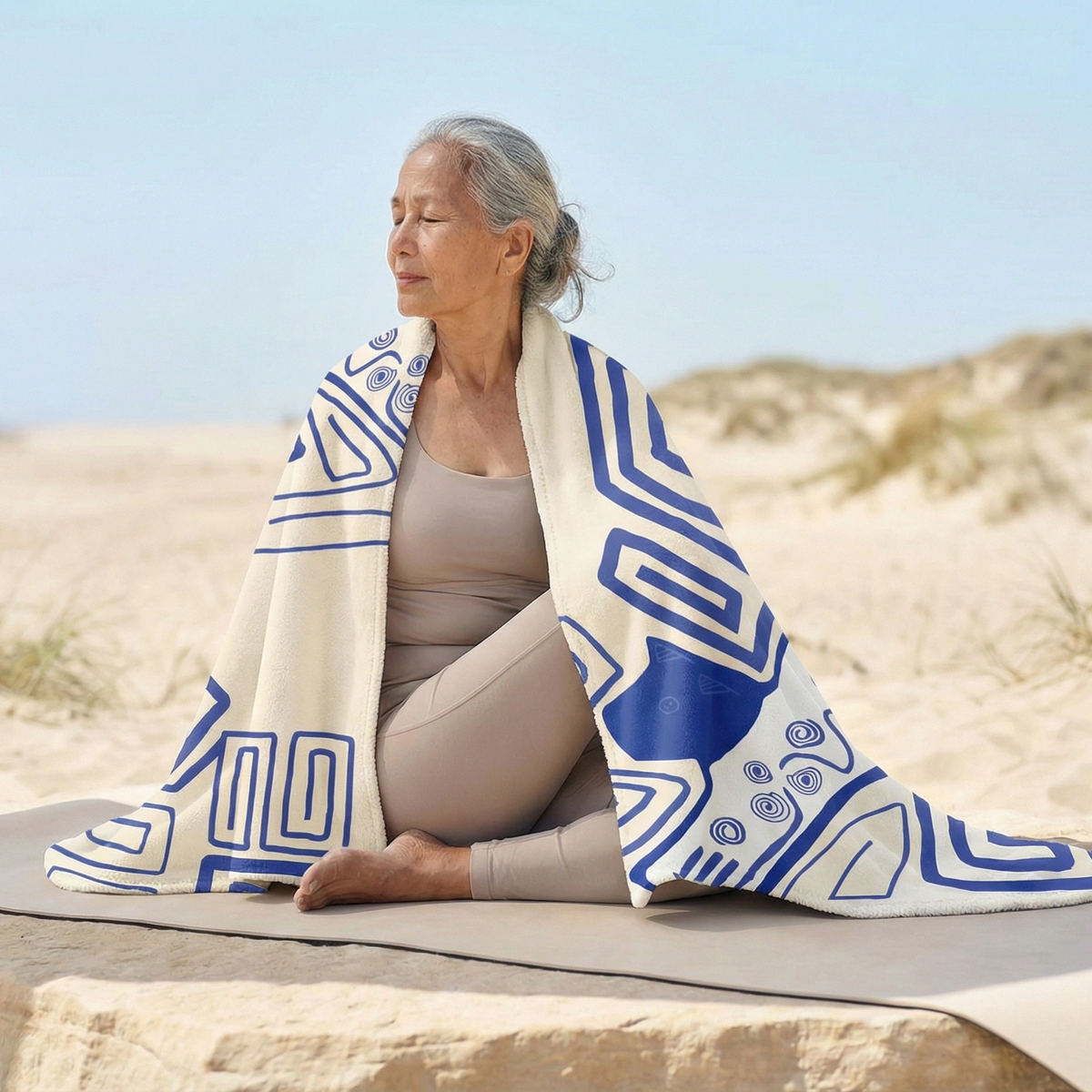 Woman seated on a stone ledge by sand dunes, wrapped in a cream Colombian Dreams blanket with blue geometric lines