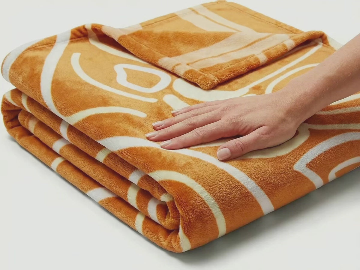 Product video: a hand smooths folded Queen of Phoenix velveteen blankets in orange, blue and grey